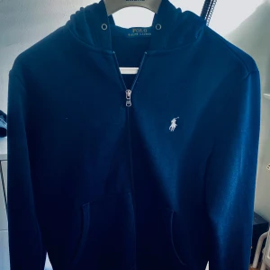 Ralph Lauren tröja - Snygg mörkblå hoodie från Polo Ralph Lauren med dragkedja och klassisk logga på bröstet. Tröjan har huva med snörningshål och två fickor framtill. Perfekt för en chill och stilren look.