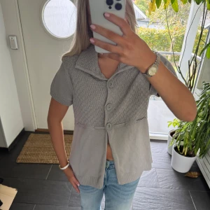 Ljusgrå stickad blus med knappar - Säljer en ljusgrå stickad blus med korta ärmar och stora knappar framtill. Toppen har en bred krage, diskret strukturmönster upptill och knytband i ryggen. Perfekt till jeans för en chill look.