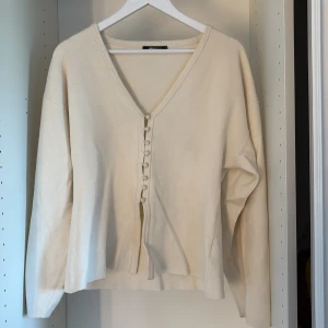 Beige kofta med knappar från Gina Tricot - Beige kofta från Gina Tricot med v-ringning och dekorativa tygklädda knappar framtill. Koftan har lång ärm och är tillverkad i ett mjukt, stickat material. Perfekt för lager-på-lager och enkel att styla till olika looks.