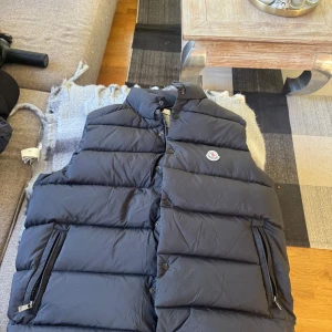 Svart dunväst från Moncler - Snygg svart dunväst från Moncler med hög krage och tryckknappar framtill. Västen har två dragkedjefickor och klassisk Moncler-logga på bröstet. Oandvänd i nytt skick 