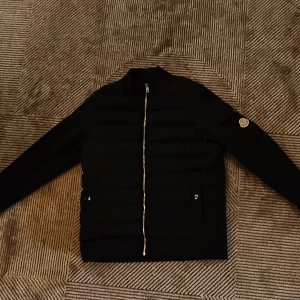 Svart pufferjacka från Moncler - Svart pufferjacka från Moncler med klassisk logga på ärmen och silvrig dragkedja framtill. Jackan har quiltad framsida, lång ärm och diskreta fickor med tryckknappar. Perfekt för dig som vill ha en clean och stilren look.