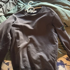 Mörkbrun sweatshirt från Stone Island - Säljer en mörkbrun sweatshirt från Stone Island med klassisk rund hals och ribbade muddar. Tröjan har den ikoniska Stone Island-loggan på vänster ärm och är tillverkad i mjuk bomull. Perfekt för dig som gillar stilrena och snygga plagg.
