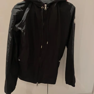 Moncler windbreaker - Svart vindjacka från Moncler med huva och dragkedja framtill. Jackan har Moncler-logga på ärmen och en snygg detalj med rödvit rand på huvan. Tillverkad i lätt polyester, perfekt för blåsiga dagar. Långärmad och stilren design.