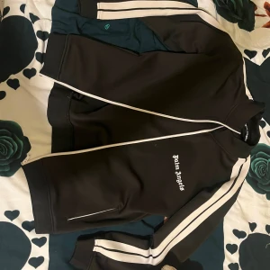 Svart track jacket från Palm Angels - Svart track jacket från Palm Angels med vita ränder längs ärmarna och vit dragkedja. Jackan har ståkrage, broderad logga på bröstet och två fickor med dragkedja. Perfekt för en sportig och trendig look.