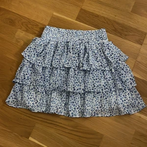 Blommig volangkjol från Sister - Supersöt kort volangkjol från Newyorker med blått blommönster på vit botten. Kjolen har flera lager volanger och bred resår i midjan för en bekväm passform. Perfekt för dig som gillar en feminin och somrig stil.