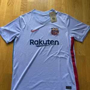 FC Barcelona bortatröja Nike M - Snygg FC Barcelona bortatröja från Nike i ljuslila polyester med klubbmärke och Nike-logga i metallic orange. Tröjan har korta ärmar, rund hals och Rakuten-tryck på bröstet. Perfekt för dig som vill visa ditt stöd för Barça på match eller träning.