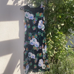 Blommig långklänning med retroskärp - Lång, ärmlös klänning med svart botten och stora blommor i vitt, blått, grönt, lila och orange. Klänningen har en retroinspirerad look och tillhörande skärp med dekorativ stor ljusgrön knapp framtill och dragkedja i ryggen. Perfekt för dig som gillar färg och mönster.