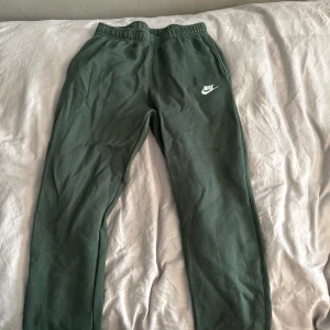 Mörkgröna mjukisbyxor från Nike - Mörkgröna mjukisbyxor från Nike med resår i midjan och vid bensluten. Byxorna har en klassisk Nike-logga i vitt på ena benet och är tillverkade i mjukt material som är perfekt för chill eller träning.