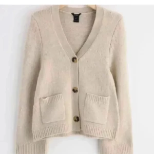 Beige kofta med fickor och knappar - Mysig beige kofta med v-ringning, tre stora knappar framtill och två praktiska fickor. Koftan har ribbade muddar och är långärmad, perfekt för lager-på-lager. Enkel och stilren design som passar till det mesta.