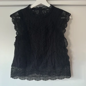 Svart spetsblus från ONLY XS - Supersnygg svart blus från ONLY i spets med högt halsringning och fina vågiga kanter. Ärmlös modell med detaljerad spets och dragkedja i ryggen. Perfekt för dig som vill ha en elegant och trendig look med extra detaljer.