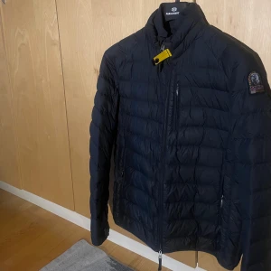 Parajumpers jacka - Säljer en svart dunjacka från Parajumpers med klassisk quiltad design och gul detalj vid kragen. Jackan har dragkedja framtill, lång ärm och en patch på axeln. Insidan är grå och jackan är lätt och smidig, perfekt för kyliga dagar.