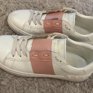 Valentino Garavani Open sneakers vit/rosa - Snygga Valentino Garavani Open sneakers i vitt skinn med bred rosa skinnpanel över snörningen. Klassisk rund tå, platt sula och diskreta nitar baktill. Guldlogga på plösen och innersulan. Perfekta för dig som vill ha en lyxig och trendig look. Inte riktiga !
