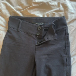 Svarta bootcut lågmidjade kostymbyxor från Gina Tricot - Svarta bootcut-byxor från Gina Tricot med två knappar och dragkedja framtill. Byxorna har fickor med lock och knapp bak samt hällor för bälte. Snygg passform och stretchigt material som känns bekvämt hela dagen.