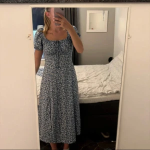 Klänning - Säljer en superfin blå långklänning med vitt blommönster och puffärmar. Klänningen har en söt snörning framtill och en smickrande passform med markerad midja. Perfekt för sommarens alla tillfällen! Materialet känns lätt och luftigt.