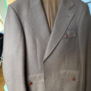 Brun ullkavaj från Strands - Säljer en brun kavaj från Strands i 100% ull. Klassisk design med breda slag, tre fickor med knappar och snygga sömdetaljer på ryggen. Perfekt för dig som gillar tidlös stil och vill ha en varm, dressad look.