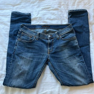 Blå jeans från Nudie Jeans, W29 L32 - Snygga blå jeans från Nudie Jeans med klassisk femficksdesign och kontrastsömmar. Jeansen har en Slim passform. Framtill samt Nudie-logga på bakfickan. Perfekt för dig som gillar stilrena och tidlösa jeans.