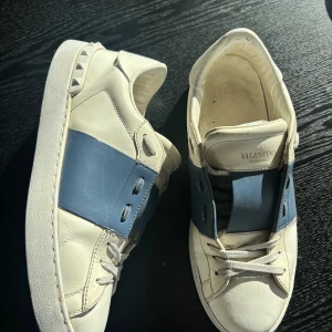 Valentino Garavani Open sneakers vit/blå - Snygga Valentino Garavani Open sneakers i vitt skinn med bred blå läderdetalj över vristen. Klassisk rund tå, platt vit sula och grå snörning. Märkeslogga på plösen och ikoniska nitar baktill. Perfekta för dig som gillar exklusiva och stilrena sneakers.