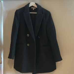 Svart tweed kappa från H&M med guldknappar - Stilren svart kappa från H&M med dubbelknäppning och snygga guldiga knappar. Jackan har en rak passform, klassisk krage och två fickor framtill. Materialet är strukturerat och ger en cool känsla. Perfekt för dig som vill ha en tidlös look. 