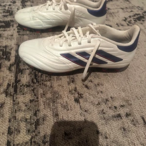 Adidas Copa Pure - vita och blå fotbollsskor - Säljer ett par Adidas fotbollsskor i vitt med blå detaljer och röda dobbar. Skorna har klassiska tre ränder på sidan, snörning och en snygg sportig design. Yttermaterialet är syntet och sulan är blå med röda dobbar för bra grepp på planen.