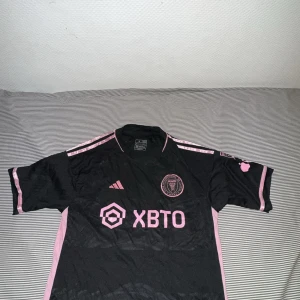 Inter Miami Messi matchtröja Adidas  - Snygg svart och rosa Inter Miami fotbollströja från Adidas med Messi 10 på ryggen. Tröjan har klubbmärke, sponsortryck och rosa detaljer längs axlarna. Tillverkad i lätt och ventilerande material, perfekt för fotboll eller att bära som supporter.