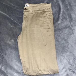 Beiga chinos från Jack & Jones - Snygga beiga chinos från Jack & Jones med klassisk passform och raka ben. Byxorna har sidfickor, bakficka med knapp och bälteshällor. Perfekta för en clean och stilren look. Materialet är mjukt och känns skönt mot huden. Storlek: 32/32. Byxan är lite skrynklig då den legat i garderoben länge. 