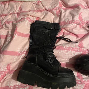 Svarta platåboots med snörning - Säljer ett par shaker 70 med rejäl platåsula och snörning hela vägen upp. Skorna har metallspänne och ringdetaljer upptill. Perfekt för dig som vill ha en edgy och cool look med extra höjd. Orginalpriset på tuffingen var 1800 kr