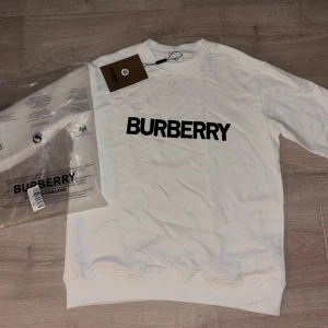 Vit sweatshirt från Burberry - Säljer en clean vit sweatshirt från Burberry med stor svart logga framtill. Tröjan är i mjuk bomull och har klassisk rund hals och långa ärmar. Perfekt för dig som gillar stilrena och exklusiva plagg med streetkänsla.