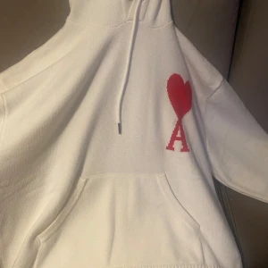 Vit hoodie från Ami Paris med rött hjärta - Vit hoodie från Ami Paris med rött hjärta och bokstaven A broderat på bröstet. Tröjan har huva med snörning och en stor magficka. Perfekt för dig som gillar stilrena och trendiga plagg med en twist.