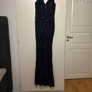 Marinblå långklänning med paljetter - Säljer en marinblå långklänning med v-ringning och ärmlös design. Klänningen är täckt av glittriga paljetter som ger en lyxig känsla. Den har en figurnära passform och är perfekt för bal eller fest. Materialet är troligtvis polyester med paljettbroderi.