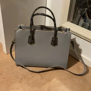 Grå axelväska med ormmönstrade handtag - Snygg grå axelväska med raka linjer och guldiga detaljer. Väskan har både axelrem och två handtag i svart ormmönstrat material. Perfekt för dig som vill ha en stilren och rymlig väska med det lilla extra.