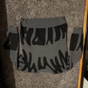 Grå och svart stickad tröja med mönster - Säljer en grå stickad tröja med svart abstrakt mönster över hela plagget. Tröjan har rund halsringning och långa ärmar. Perfekt för dig som gillar en unik och cool stil med grafiska detaljer.