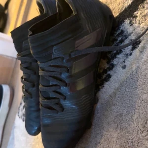 Svarta Adidas fotbollsskor - Säljer ett par svarta Adidas fotbollsskor med klassiska tre ränder på sidan. Skorna har snörning och är tillverkade i syntetmaterial med en modern, sportig design. Perfekta för dig som vill ha stilrena och funktionella skor på planen.