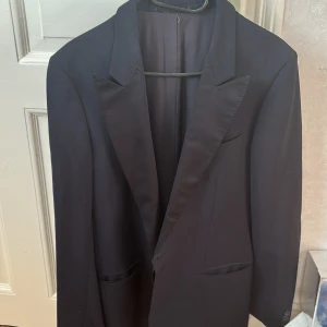 Svart kavaj från Atelier Torino - Säljer en klassisk svart kavaj från Atelier Torino, specialtillverkad för Holzherr. Kavajen är tillverkad i Italien av 65% ull och 35% mohair, med foder i viskos. Den har spetsiga slag, två fickor fram och en bröstficka. Perfekt för dig som vill ha en stilren och tidlös look.