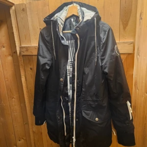 Svart parkajacka från bpc med huva - Svart parkajacka från bpc med vit dragkedja, stora fickor och justerbar huva. Jackan har ett rutigt innerfoder i beige, svart och grått. Snygga detaljer och patch på ärmen. Perfekt för kyliga dagar.