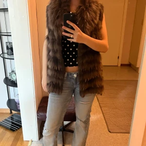 Brun lång pälsväst med fluffig krage - Säljer en brun lång pälsväst med bred, fluffig krage och horisontella ränder i pälsen. Västen är ärmlös och har en loose passform, perfekt att slänga över en hoodie eller tröja för extra stilpoäng.