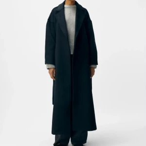 Svart lång kappa med bälte - Stilren svart kappa i lång modell med bred krage och knytbälte i midjan. Kappan har en rak och loose passform, långa ärmar och är tillverkad i ett mjukt ullmaterial. Perfekt för dig som gillar minimalistisk och clean stil. Normal pris 1499.
