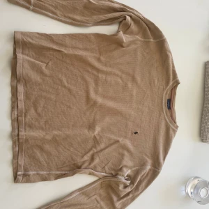 Beige långärmad tröja Polo Ralph Lauren - Beige långärmad tröja från Polo Ralph Lauren i 100% bomull. Tröjan har en diskret broderad logga på bröstet och är tillverkad i ett ribbat, mjukt material. Perfekt för en chill och stilren look.