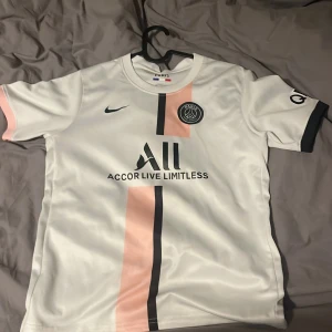 PSG fotbollströja Nike vit/rosa - Paris Saint-Germain fotbollströja i vitt med ljusrosa och svart vertikal rand. PSG-logga på bröstet och Nike Swoosh på andra sidan. Tillverkad i lätt och ventilerande material, perfekt för match eller träning. Klassisk rund hals och korta ärmar. Med neymar jr på ryggen men det har gått bort.