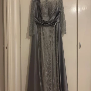 Lång glittrig silverklänning med mesh - Snygg långklänning i silver med glittriga detaljer och transparent mesh upptill. Klänningen har långa ärmar, markerad midja och elegant fall. Dragkedja bak för enkel påklädning. Perfekt för dig som vill sticka ut med en glamorös look. 