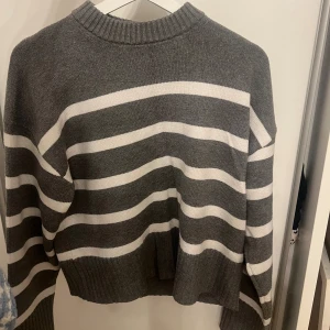 Randig gråvit stickad tröja - Säljer en stickad tröja i grått och vitt med breda horisontella ränder. Tröjan har ribbad halsringning, muddar och nederkant samt lång ärm. Perfekt för dig som gillar en avslappnad och stilren look.
