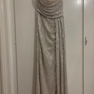Glittrig långklänning i champagne - Säljer en maxilång klänning i champagnefärgat tyg med glittrande paljetter över hela ytan. Klänningen har draperad överdel, smala axelband och en snygg omlottkänsla framtill. Perfekt för dig som vill sticka ut med en elegant och lyxig look. Dragkedjan är sönder men man kan fixa den, jag köpte den för 2500 kr och har använt den bara en gång den är jättebra stick men bara att dragkedjan är sönder 