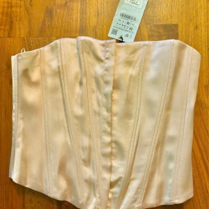 Champagnefärgad korsett från Zara - Säljer en snygg champagnefärgad korsett från Zara i glansig polyester. Toppen är axelbandslös och har markerade sömmar framtill för en strukturerad look. Perfekt att styla med jeans eller kjol för en trendig outfit.