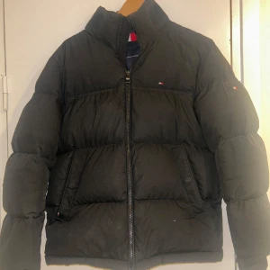 Tommy hilfiger jacka - Säljer en svart pufferjacka från Tommy Hilfiger med hög krage och dragkedja framtill. Jackan har diskret logga på bröstet och ärmen, samt två fickor på sidorna. Perfekt för kalla dagar och riktigt snygg till streetwear-stilen. Nypris ca 2000kr