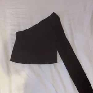 Svart oneshoulder topp - Säljer en svart oneshoulder topp med lång ärm. Toppen är croppad och har en tight passform som sitter snyggt på kroppen. Perfekt för dig som vill ha en stilren och trendig look. Materialet är mjukt och stretchigt för extra komfort.