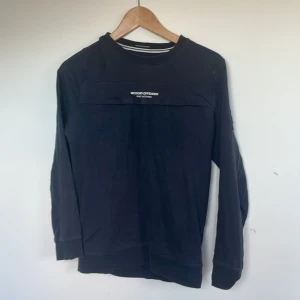 Svart långärmad tröja Weekend Offender XS - Svart långärmad tröja från Weekend Offender i 100% bomull. Tröjan har en cool detalj med ficka och logga framtill samt patch på ärmen. Perfekt för dig som gillar streetwear och enkel stil.