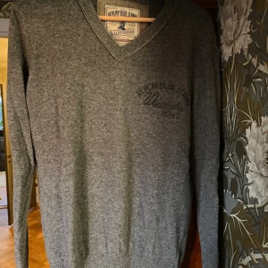 Grå v-ringad tröja Replay Blue Jeans - Grå v-ringad tröja från Replay Blue Jeans med diskret broderad logga på bröstet. Tröjan har lång ärm och är tillverkad i mjukt material som känns skönt mot huden. Perfekt för lager-på-lager och enkel att matcha med jeans eller chinos.