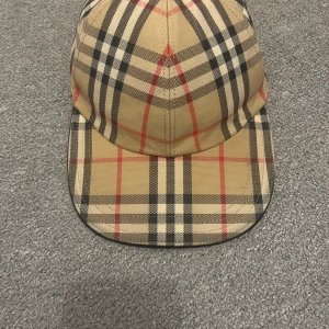 Burberry rutig keps den är äkta - Snygg keps från Burberry i klassiskt beige rutigt mönster med svarta, vita och röda detaljer. Justerbart spänne baktill i metall med Burberry-logga. Tillverkad i bomull och har en böjd skärm. Perfekt accessoar för en trendig look.