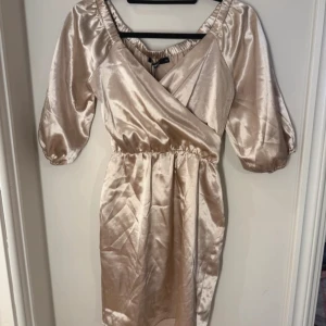 Champagnefärgad omlottklänning från BikBok - Säljer en elegant champagnefärgad omlottklänning från BikBok. Klänningen har en offshoulder-design med trekvartsärmar och är tillverkad i ett glansigt satinmaterial. Perfekt för en stilfull look! Säljer för att den är lite för stor för mig som vanligtvis bär xs/s