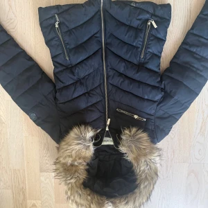 Svart pufferjacka Boohoo med fuskpäls - Svart pufferjacka från Boohoo med snygga guldfärgade dragkedjor och huva med fuskpäls. Jackan har quiltad design och insida i leopardmönster som ger extra edge. Perfekt för kalla dagar när du vill sticka ut lite extra.