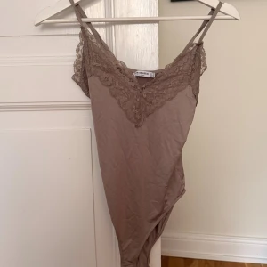 Beige linnebody med spets från Stradivarius - Supersnygg beige linnetopp (body) från Stradivarius med tunna axelband och v-ringning dekorerad med spets. Mjuk och stretchig i materialet, perfekt att styla med jeans eller kjol. Justerbara axelband för bästa passform.